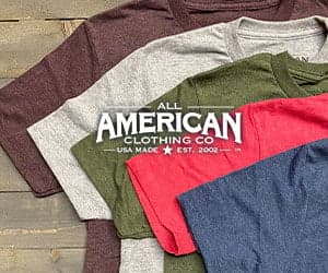 Folded t-shirts in several colors on a wood surface, with the All American Clothing Co. logo overlay, from official promotional imagery.
